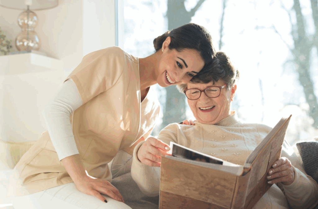 An older adult with dementia laughs while showing a caregiver a photo album in front of a sunny window in memory care
